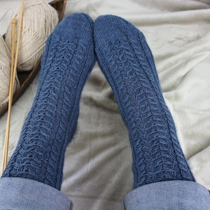 May include: A pair of blue knitted socks featuring a vertical cable knit pattern running down the centre of the foot and leg. The socks are crafted from a textured yarn and are shown extending from the cuffs of blue denim jeans.