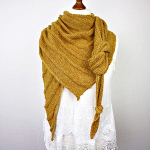 Könnte beinhalten: Senfgelber Strickschal mit einem strukturierten, diagonal gerippten Muster. Der Schal ist über einer weißen Schneiderpuppe drapiert und vorne verknotet, mit langen, geknoteten Enden, die herunterhängen. Der Schal hat eine dreieckige Form.