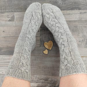 Könnte beinhalten: Ein Paar hellgraue Zopfstricksocken. Die Socken haben ein strukturiertes Muster und werden auf einer Holzoberfläche gezeigt. Zwei kleine Holzherzformen sind ebenfalls sichtbar.