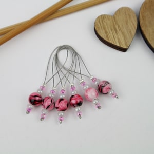 Könnte beinhalten: Satz mit sechs rosafarbenen und schwarzen marmorierten Perlen-Maschenmarkierern mit silbernen Drahtschlaufen. Die Perlen sind mit kleinen rosafarbenen und weißen Perlen verziert.