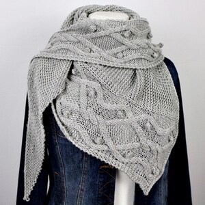 Könnte beinhalten: Ein hellgrauer Strickschal mit einem strukturierten Zopfmuster und Bommeldetails. Der Schal ist über einer Jeansjacke drapiert und zeigt seine dreieckige Form und die komplizierten Muster. Dieses gemütliche Accessoire ist perfekt, um Wärme und Stil zu verleihen.