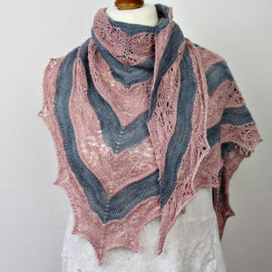 Puede incluir: Un chal tejido a mano con un diseño de punto calado. El chal presenta rayas alternas en rosa empolvado y gris azulado. Los bordes tienen un acabado festoneado y puntiagudo. El chal está drapeado sobre un maniquí blanco.