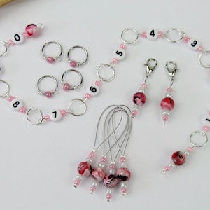 Könnte beinhalten: Ein Set aus rosa und weißen Maschenmarkierern mit den Zahlen 0-9, eine Kette aus Maschenmarkierern mit den Zahlen 1-5 und vier Nadelhalter mit rosa und weißen Perlen.