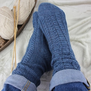 Könnte beinhalten: Nahaufnahme von blauen Strickstrümpfen mit Zopfmuster. Die Socken sind über den Knöcheln umgeschlagen und werden mit einer blauen Jeans getragen. Im Hintergrund befinden sich Wollknäuel und Stricknadeln.