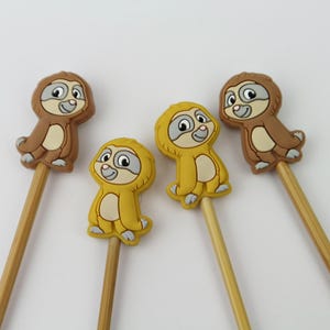 Könnte beinhalten: Vier Holzstifte mit aufgesetzten Faultierfiguren aus Gummi. Zwei Faultiere sind braun mit cremefarbenem Bauch und Gesicht, während die anderen beiden gelb mit cremefarbenem Bauch und Gesicht sind. Jedes Faultier hat große Augen und eine kleine Nase.