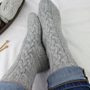 Könnte beinhalten: Ein Paar hellgraue Strickstrümpfe mit einem vertikalen Zopfmuster, das über den Fuß und das Bein verläuft. Die Socken haben ein geripptes Bündchen am oberen Rand und werden zu einer blauen Jeans getragen, was die strukturierte Wolloptik betont.
