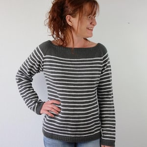 Könnte beinhalten: Ein grau-weiß gestreifter Pullover mit U-Boot-Ausschnitt. Die langärmelige Strickware hat horizontale Streifen und einen gerippten Saum. Der Pullover ist ein klassisches Design, das sich für Freizeitkleidung eignet. Das Model trägt eine blaue Jeans.