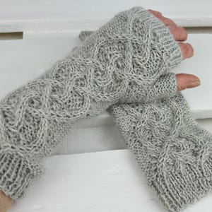 Könnte beinhalten: Ein Paar hellgraue Fäustlinge mit Zopfmuster. Die Handschuhe sind aus weichem, strukturiertem Garn gefertigt und weisen ein detailliertes Zopfmuster auf. Die Handschuhe sind so konzipiert, dass sie die Hände warm halten und gleichzeitig die Finger frei lassen.