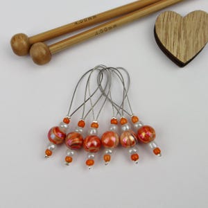 Könnte beinhalten: Eine Sammlung von Maschenmarkierern mit orangefarbenen und roten Glasperlen und silbernen Drahtschlaufen. Zwei Stricknadeln aus Holz, markiert mit "4,00 mm", und ein Holzherz sind ebenfalls vorhanden.