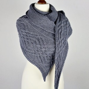 May include: A gray knitted shawl with a textured cable knit design. The triangular shawl is draped over a mannequin, showcasing the intricate pattern and soft texture of the yarn. The shawl is a muted gray color.