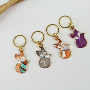 Könnte beinhalten: Vier goldene Maschenmarkierer mit Fuchs-Anhängern. Die Füchse tragen verschiedene farbige Outfits. Ein Fuchs ist lila, einer ist orange und weiß, einer ist grau und weiß und einer ist orange und blau.