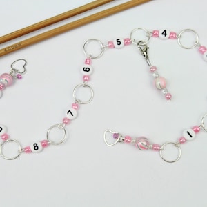 Könnte beinhalten: Zwei Sätze rosa und silberner Maschenmarkierer mit den Zahlen 0-9 und 1-4. Die Markierer sind an einer Kette mit einem Herz-Anhänger und einer rosa-weißen Perle befestigt.