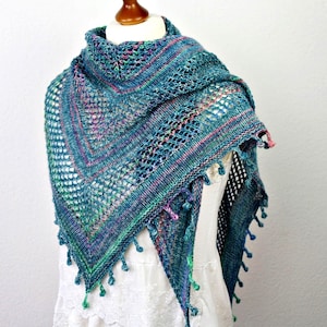 Puede incluir: Un chal tejido de color verde azulado y azul, de forma triangular, exhibido en un maniquí. El chal presenta un delicado diseño calado, con detalles en rosa y verde. Pequeñas borlas adornan los bordes.