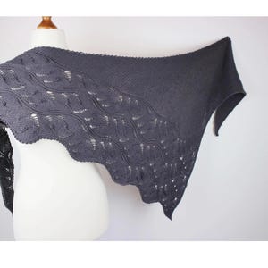 Puede incluir: Un chal triangular tejido en color gris oscuro que presenta un patrón de ondas texturizado en el borde inferior y una sección de punto liso en la parte superior. El chal está drapeado sobre un maniquí blanco, mostrando su diseño de encaje.
