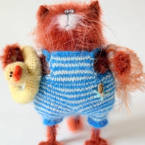 May include: A fuzzy, rust-coloured needle-felted cat figurine wearing blue and white striped knit overalls. The cat holds a small yellow duck-shaped floatie in one paw and features a small blue starfish charm attached to the front of the overalls.