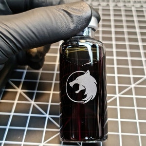 Może przedstawiać: Mała, przezroczysta butelka szklana z wygrawerowanym na przodzie czarno-białym logo wilka. Butelka jest wypełniona ciemnym płynem.