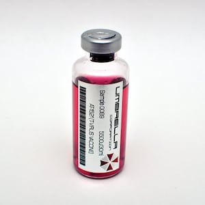 Op de afbeelding: Een heldere glazen flacon met een zilveren dop, gevuld met een helderrode vloeistof. De flacon heeft een etiket met de tekst "UMBRELLA CORPORATION" en een streepjescode. De flacon is gelabeld met "Sample 0089" en "5500J/30ml".