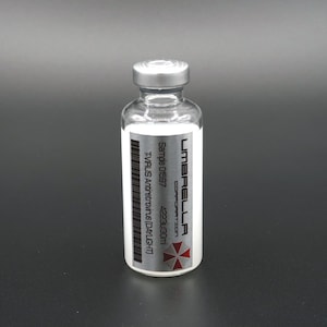 Peut inclure: Un flacon en verre transparent avec un bouchon argenté et une étiquette blanche. L'étiquette porte le texte "UMBRELLA CORPORATION" et un code-barres. Le flacon contient une substance blanche.