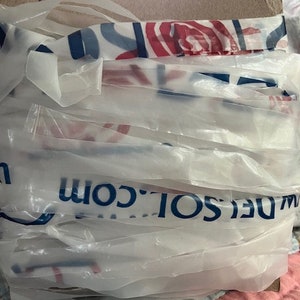 Puede incluir: Una pila de bolsas de plástico transparentes, algunas con un estampado floral azul y rojo, y el texto "W.DELSOL.COM" impreso en ellas. Las bolsas están envueltas alrededor de una base de cartón, con el texto manuscrito "27yd" en la parte superior.