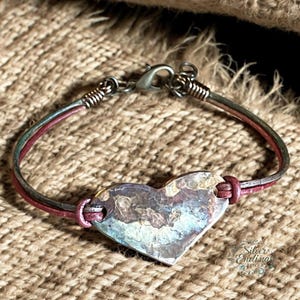 May include: A handmade bracelet featuring a heart-shaped pendant. The pendant is a mix of silver, brown, and blue hues. The bracelet has a silver clasp and is made with red and green leather strands.