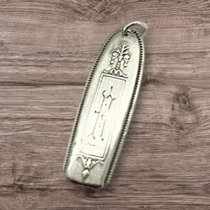 May include: A silver-colored, elongated pendant with ornate engraved details. The pendant features a decorative top loop and a rectangular central design with a stylized floral motif. The edges are lined with a beaded pattern.