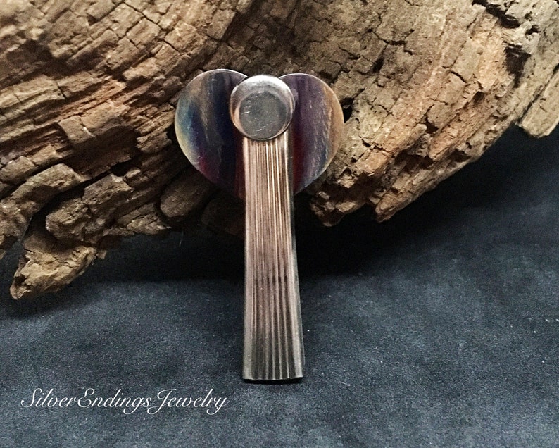 May include: A silver angel pendant with a heart-shaped top and a textured body. The heart is iridescent with shades of purple, blue, and green.