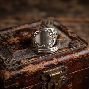 May include: A silver-toned ring crafted from a repurposed spoon handle, featuring an embossed floral pattern on the bowl section and a smooth, curved band that wraps around to form a circular shape with a polished metallic finish.