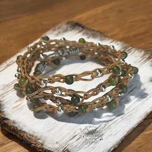 May include: A wrap bracelet featuring woven tan leather strands, interspersed with small, round, silver beads and green beads. The bracelet is displayed on a white and brown wooden surface. The bracelet is from Silver Endings Jewelry.