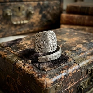 May include: A silver-toned wrap ring featuring an embossed floral pattern across the wide band. The metal surface displays intricate details of petals and small buds, with an open-ended design that curves around to form a continuous decorative motif.