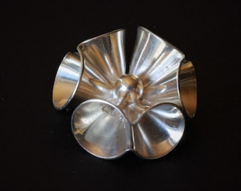 May include: A metallic silver-toned object featuring five flared, trumpet-shaped segments arranged in a circular, flower-like pattern around a central spherical core. The surface has a smooth, reflective finish with visible light reflections.