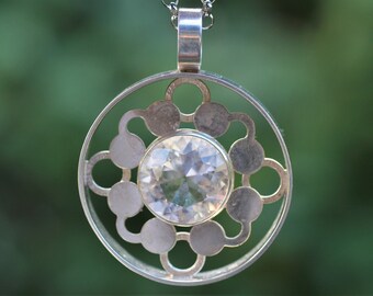 May include: A circular silver pendant featuring a central faceted clear gemstone surrounded by a geometric pattern of interconnected silver circles and discs, suspended from a silver chain with a polished metal bail at the top.