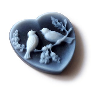 May include: A blue heart-shaped cameo featuring two white birds perched on a branch. The cameo is made from a smooth, polished material.