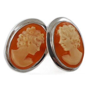 May include: A pair of silver cameo earrings with orange and white cameo insets. The cameos feature a profile of a woman's face.