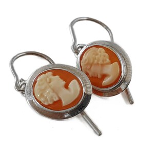 May include: A pair of silver-toned earrings featuring oval cameo designs. Each earring has a light orange background with a white profile of a woman. The earrings have a hook closure.
