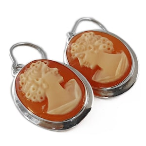 May include: A pair of oval cameo earrings. Each earring features a profile of a woman carved in cream-colored relief against an orange background, set in a silver-toned frame. The earrings have a simple hook design.
