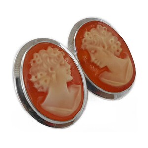 May include: Oval-shaped cameo earrings featuring a profile of a woman's face carved in orange and cream tones. The cameo is set in a silver-colored metal frame. The earrings are displayed against a white background.