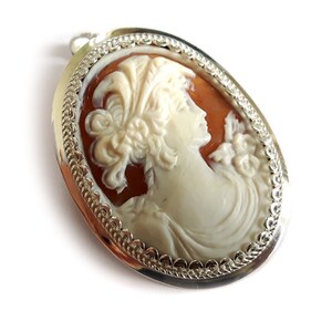 May include: A silver cameo pendant with a white cameo of a woman with a floral headdress. The cameo is set in a silver oval frame with a decorative border.