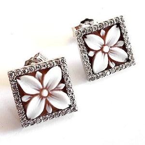May include: A pair of silver earrings with a square design. Each earring features a white cameo carved with a flower design, surrounded by a border of clear cubic zirconia stones.