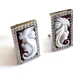 May include: A pair of silver earrings with a cameo design. Each earring features a white cameo of a woman's face set in a silver frame with clear cubic zirconia stones.