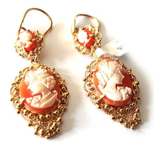 May include: A pair of gold earrings with a cameo design. Each earring features a delicate filigree design and a cameo of a woman's profile. The cameos are a light orange color with a white cameo design.