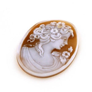 May include: A cameo carved from shell, featuring a woman with a floral crown and a delicate flower in her hand. The cameo is set in a brown oval frame.