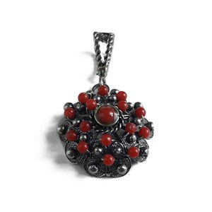 May include: A silver pendant with a red coral centerpiece and smaller red coral beads surrounding it. The pendant is in the shape of a flower with a detailed design.