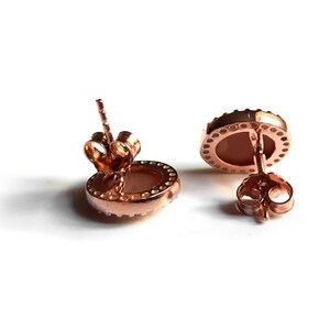 May include: A pair of rose gold stud earrings with a round, light pink stone set in a bezel setting. The earrings have a simple, elegant design.