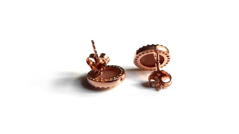 May include: A pair of rose gold stud earrings with a round, light pink stone set in a bezel setting. The earrings have a simple, elegant design.