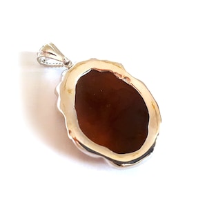 May include: A silver pendant with a brown oval-shaped stone set in a decorative silver frame. The back of the pendant is visible, showing the smooth surface of the stone.