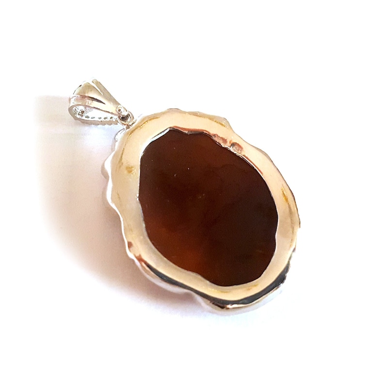 May include: A silver pendant with a brown oval-shaped stone set in a decorative silver frame. The back of the pendant is visible, showing the smooth surface of the stone.
