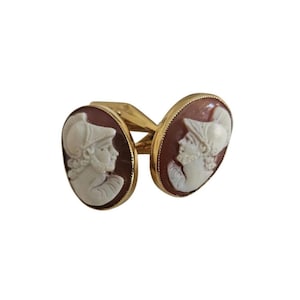 May include: A pair of gold cufflinks featuring oval cameo portraits of a man in profile. The cameos are white against a reddish-brown background, set in gold frames. The cufflinks are connected by a gold bar.