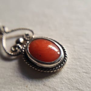 May include: A silver pendant with a heart-shaped bail and an oval-shaped coral stone set in a silver bezel. The pendant has a twisted rope design around the edge of the bezel.
