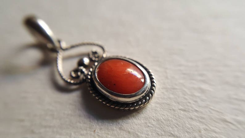 May include: A silver pendant with a heart-shaped bail and an oval-shaped coral stone set in a silver bezel. The pendant has a twisted rope design around the edge of the bezel.