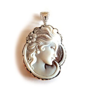 May include: A cameo pendant featuring a carved portrait of a woman with a flower in her hair. The cameo is set in a silver-tone frame with a scalloped edge.